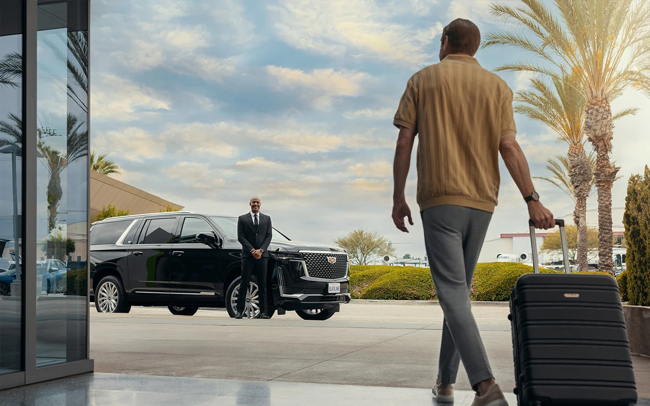 Man arriving at a luxury car service with a suitcase, greeted by a chauffeur standing beside a black Cadillac SUV, with palm trees and modern architecture in the background.