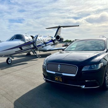 Black Lincoln sedan with "SNKS 01" plate parked near a small private jet on an airport tarmac.
