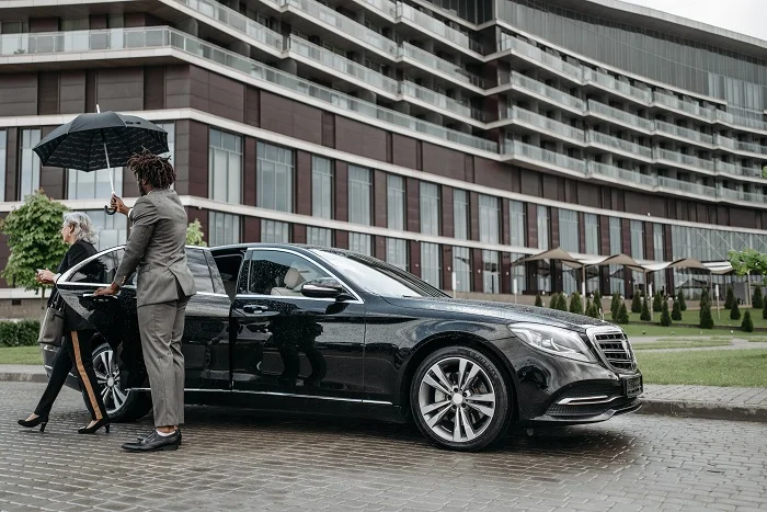 Chauffeur holding an umbrella for a woman as she exits a luxury black car in front of a hotel, representing professional NYC limo service.