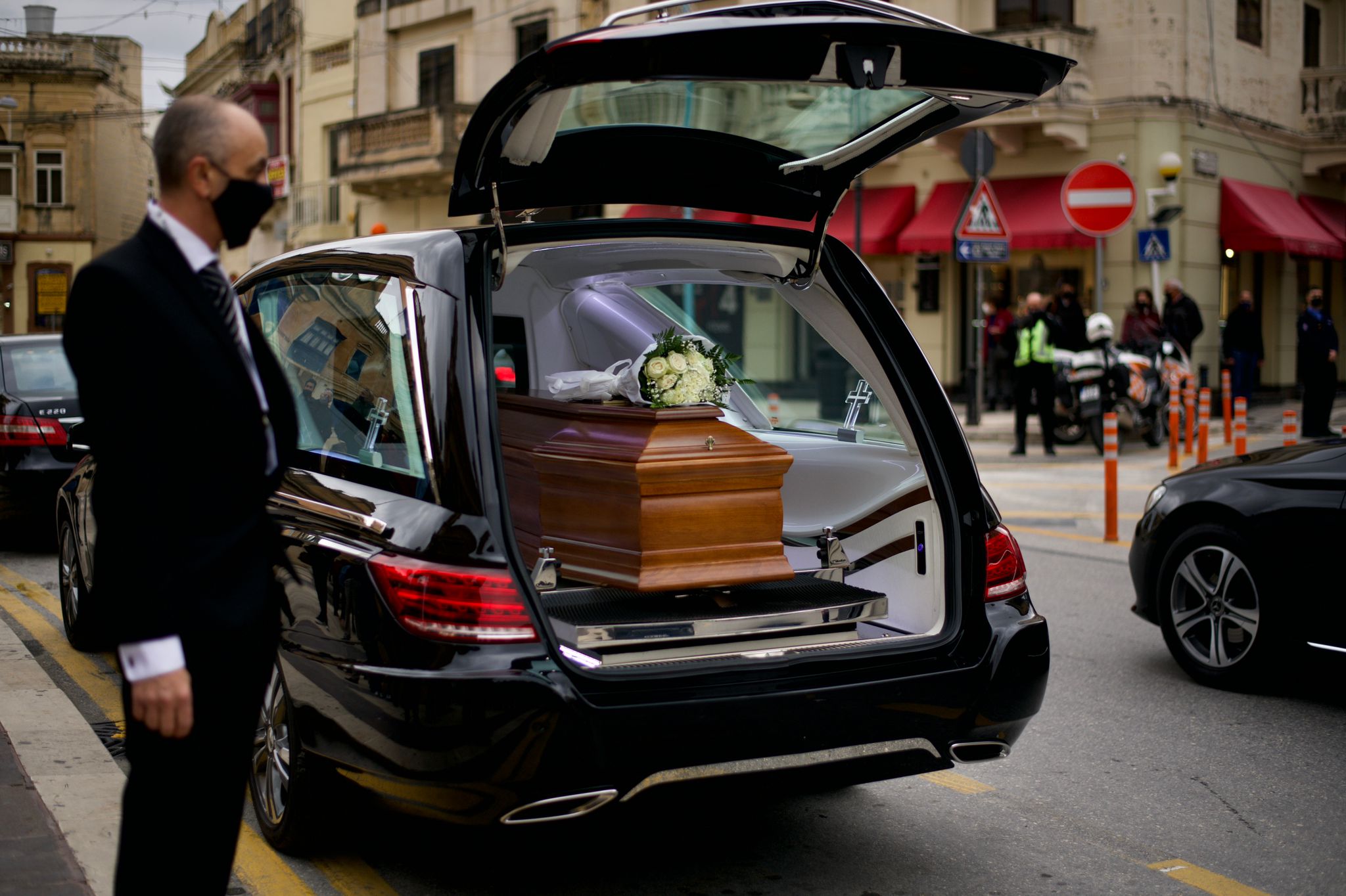 A black funeral hearse parked on a city street with an open trunk, displaying a wooden casket adorned with white flowers.