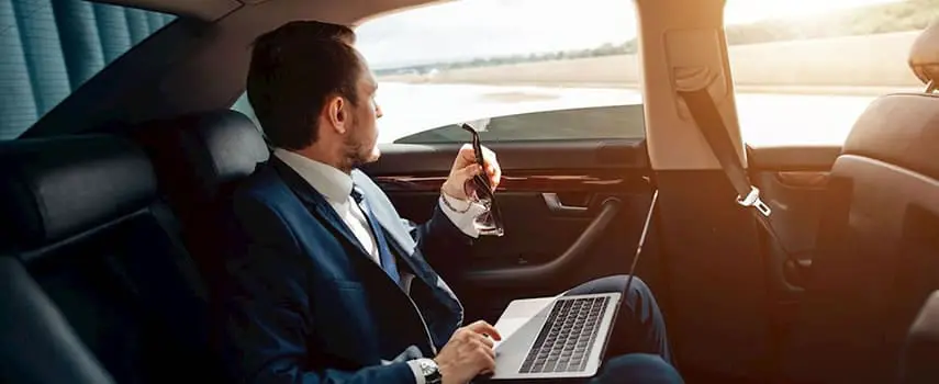 Businessman in a luxury car rental, working on a laptop while enjoying a comfortable ride, symbolizing premium travel.